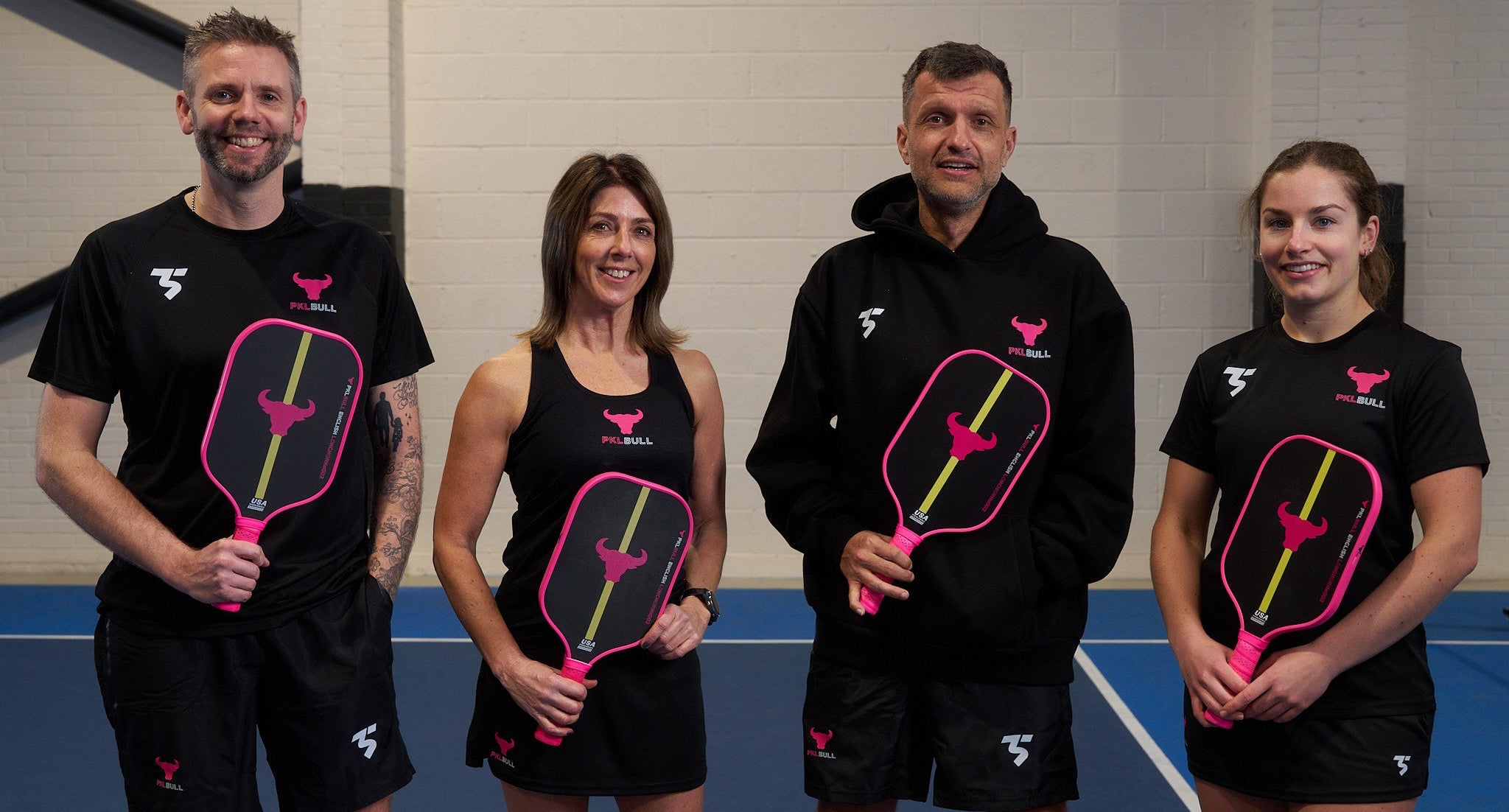 Four people standing on a pickleball court holding PKLBull paddles, wearing PKLBull branded sports attire.